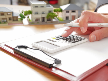 Vers la page Estimation. Une main utilise une calculatrice sur un bloc-notes, avec des maquettes de maison.