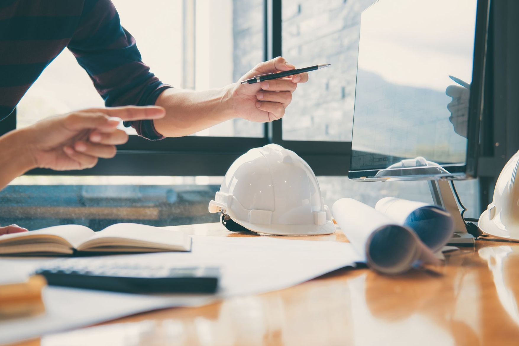 Un architecte désigne un écran d'ordinateur affichant des plans, un casque de chantier blanc est posé sur le bureau.